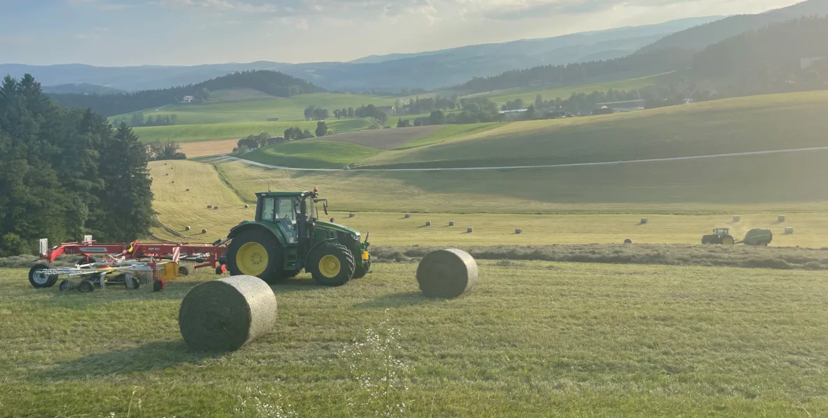 grüner Traktor und Heuballen auf Acker bei der Heuernte im Sonnenschein